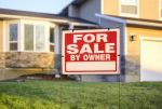 A close up of a For Sale By Owner sign, with a two-story house in the out of focus background.